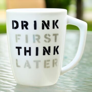 White ceramic mug on a glass table with the phrase “Drink First Think Later” stenciled in bold black and gray letters.