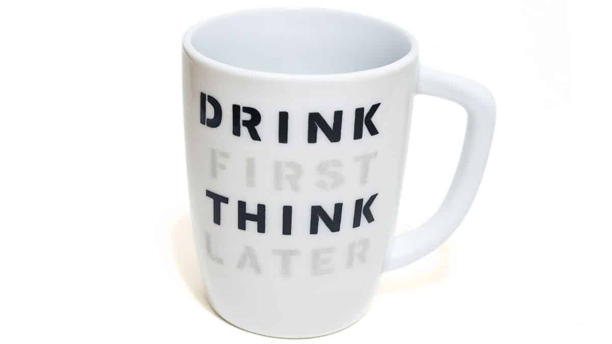 White ceramic coffee mug with the phrase “Drink First Think Later” stenciled in bold black and gray letters on the front.