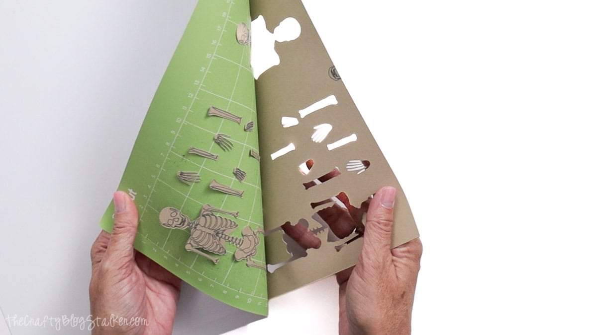 Hands peel kraft cardstock from a Cricut cutting mat to reveal cut skeleton pieces for a Halloween banner.