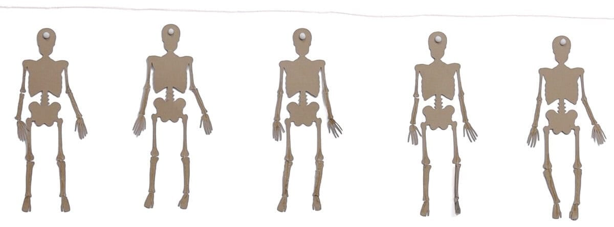 Back of five kraft paper skeletons with hanging holes arranged beneath a length of string for a Halloween skeleton banner.