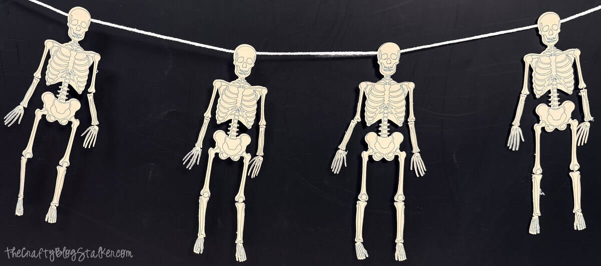 Three paper skeletons hang from a white cord against a black background as a Halloween skeleton banner.