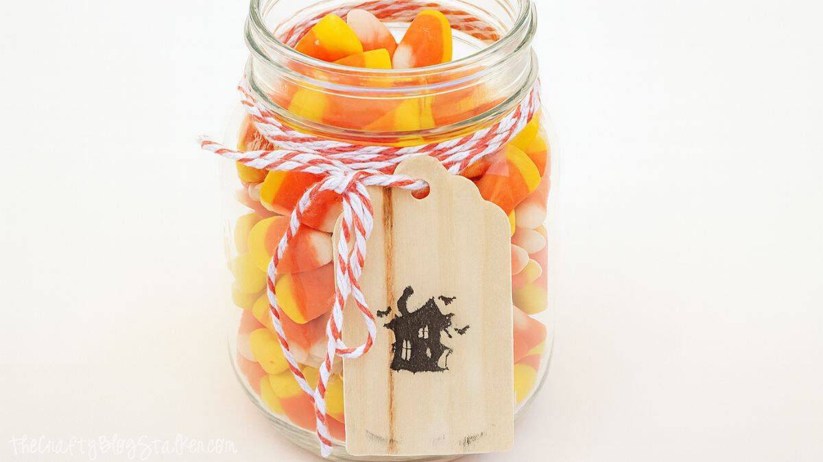 Glass jar filled with candy corn, tied with orange and white twine, and decorated with a wooden tag featuring a black haunted house design.