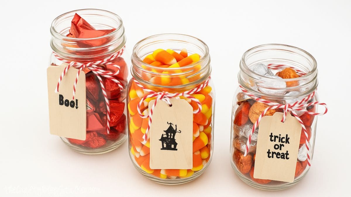 Three mason jars filled with Halloween candy, each tied with red and white twine and wooden tags labeled Boo!, a haunted house, and trick or treat.