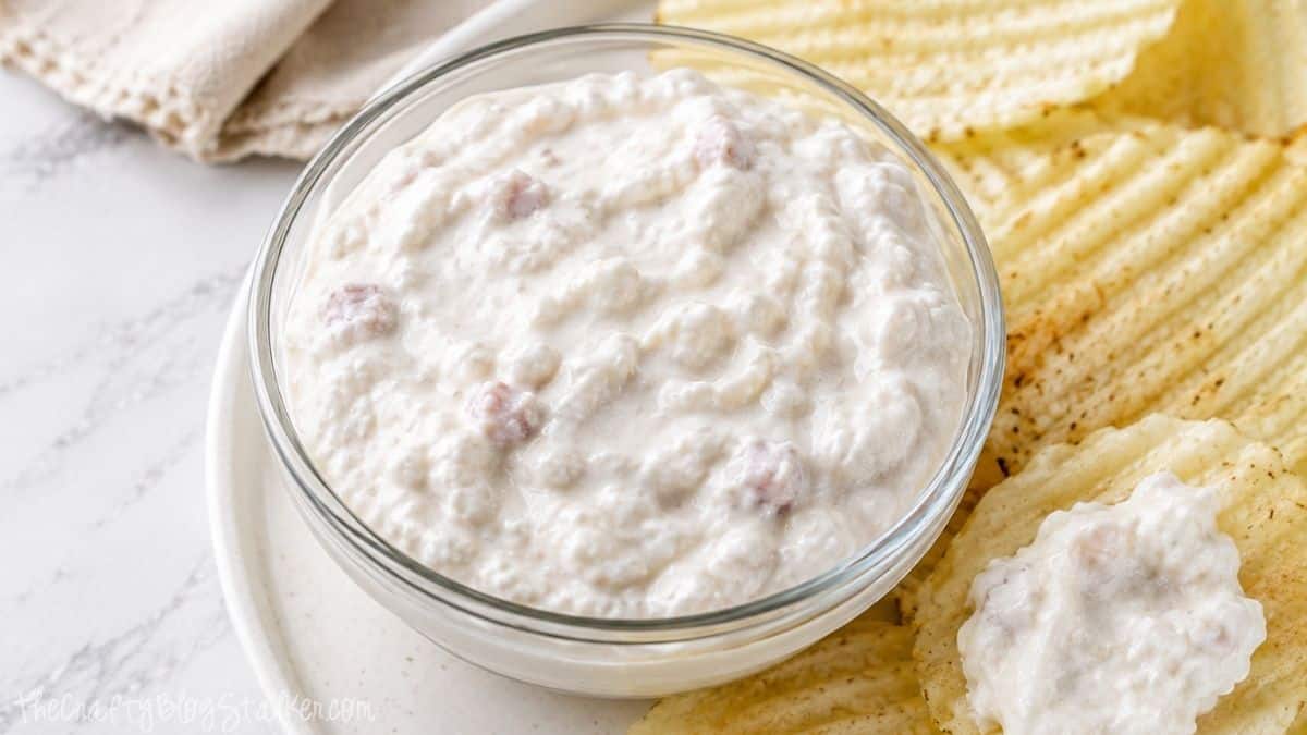 Creamy clam dip with cottage cheese served in a glass bowl with ridged potato chips on a white plate.