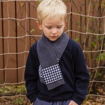 A young boy wearing a dark gray fleece scarf with a black-and-white checkered pocket, standing outdoors in front of a netted fence.