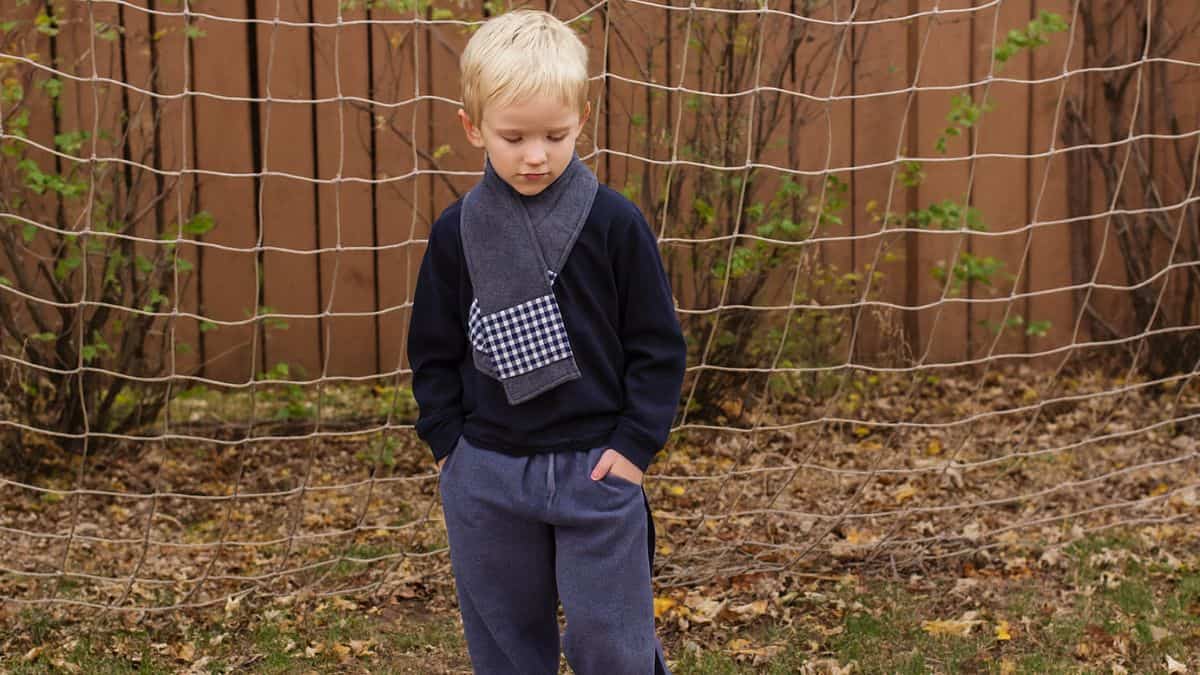 Young boy standing outdoors in front of a soccer net, wearing a dark sweatshirt, sweatpants, and a fleece scarf with a plaid pocket detail.