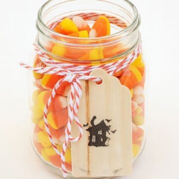 Glass jar filled with candy corn, tied with orange and white twine, and decorated with a wooden tag featuring a black haunted house design.