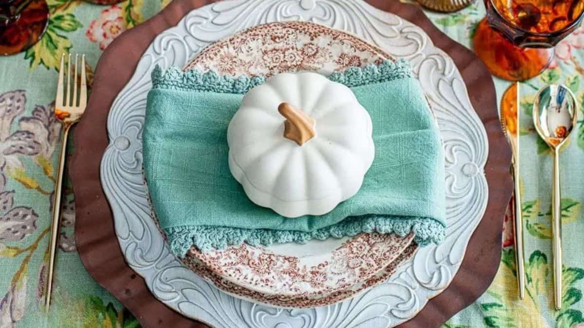 Thanksgiving place setting with layered plates, aqua napkin, gold flatware, and a small white pumpkin centerpiece on a floral tablecloth.