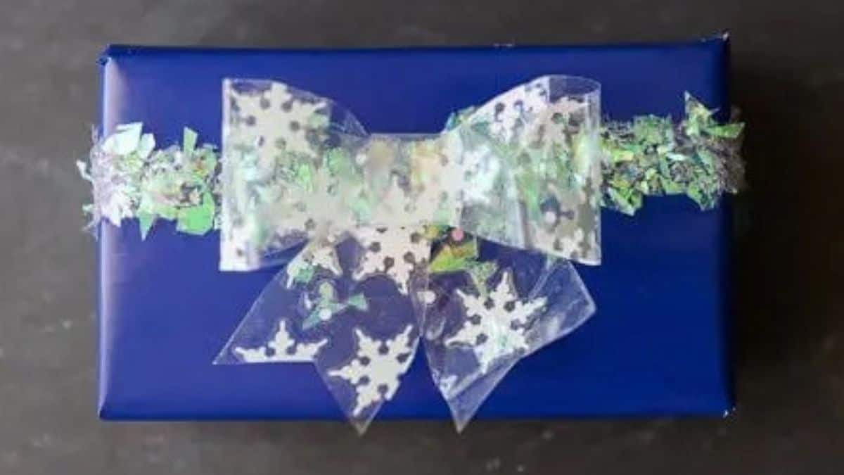 Blue wrapped gift decorated with a handmade clear bow featuring white snowflakes and iridescent shredded accents.