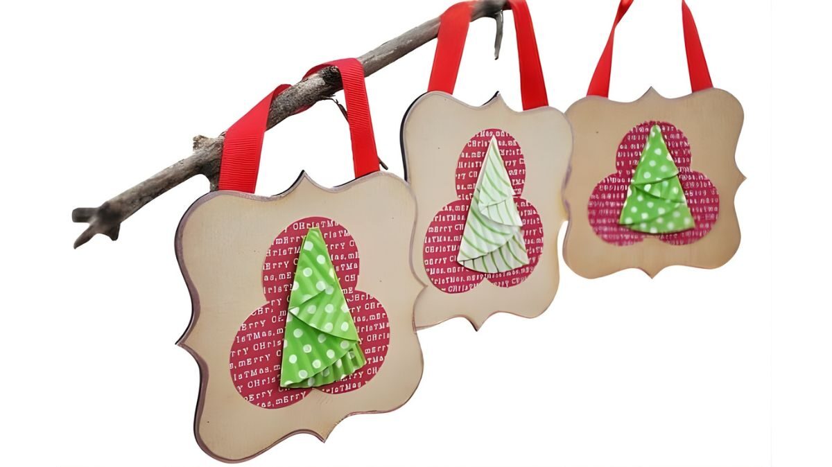 Three handmade Christmas ornaments with layered cupcake-liner trees hang from a branch on decorative wood plaques with red ribbon loops.
