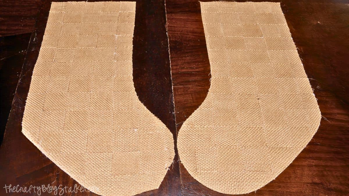 Weaved Burlap Christmas Stockings Tutorial Two burlap stocking pieces cut into matching shapes, laid flat side by side on a wooden surface, ready to be assembled into a DIY Christmas stocking.