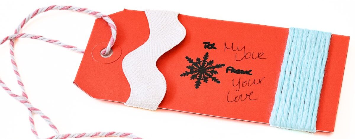 Handmade red gift tag decorated with white trim, blue wrapped twine, a snowflake stamp, and handwritten to-and-from notes, attached to striped twine.