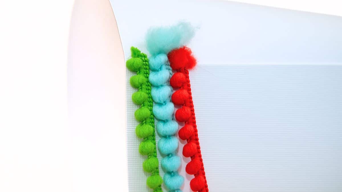 Close-up of green, blue, and red pom pom trim hot glued to the edge of a white paper cone for a craft project.