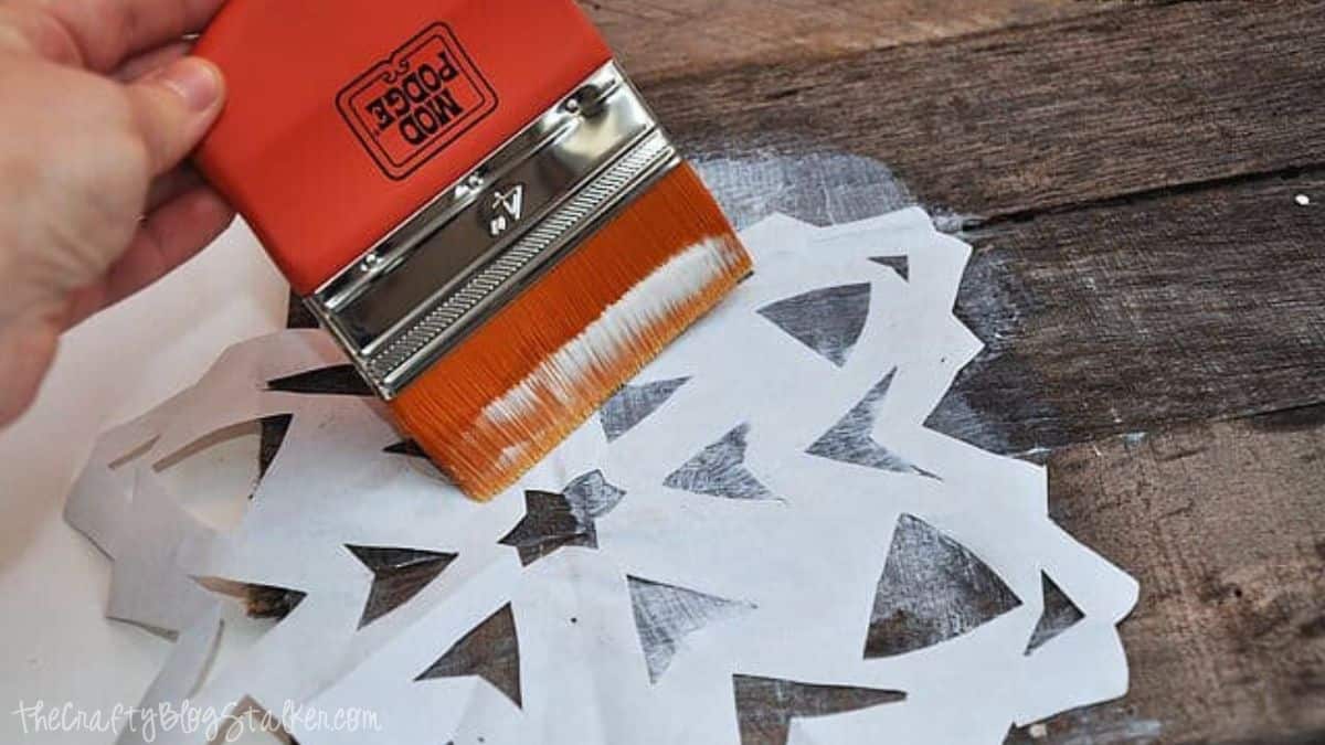 Hand using a wide Mod Podge brush to apply adhesive over a white paper snowflake stencil on a rustic wooden board for a DIY holiday craft project.