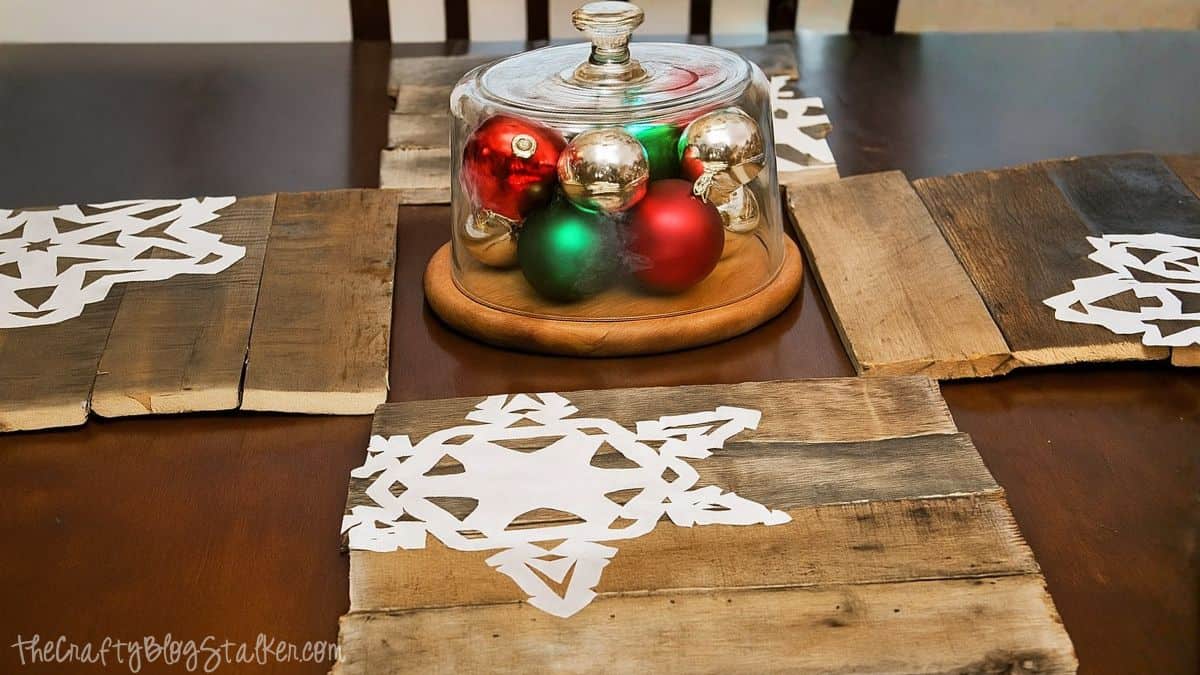 Rustic holiday table with wooden pallet chargers featuring white snowflake designs and a glass cloche centerpiece filled with red, green, and gold ornaments.
