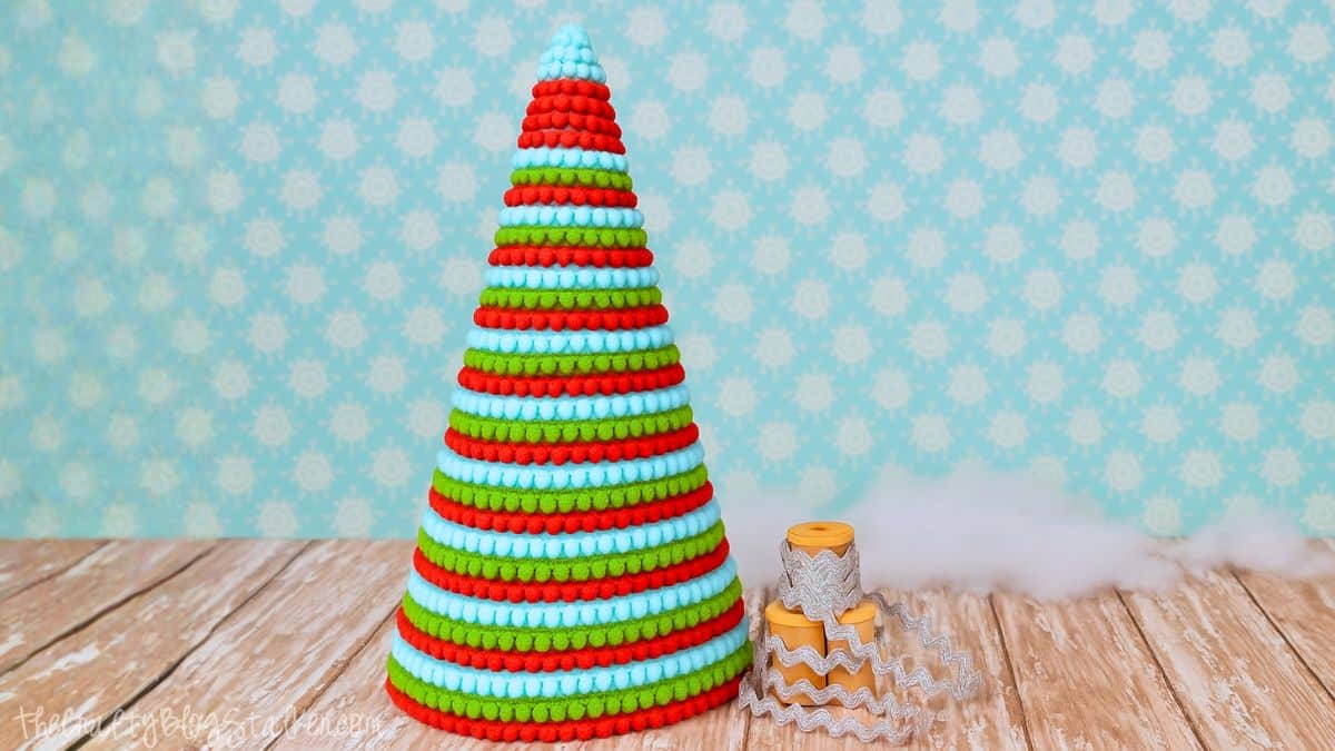 Colorful cone-shaped Christmas tree made with red, green, and blue pom pom trim, displayed on a wooden surface with ribbon spools beside it.