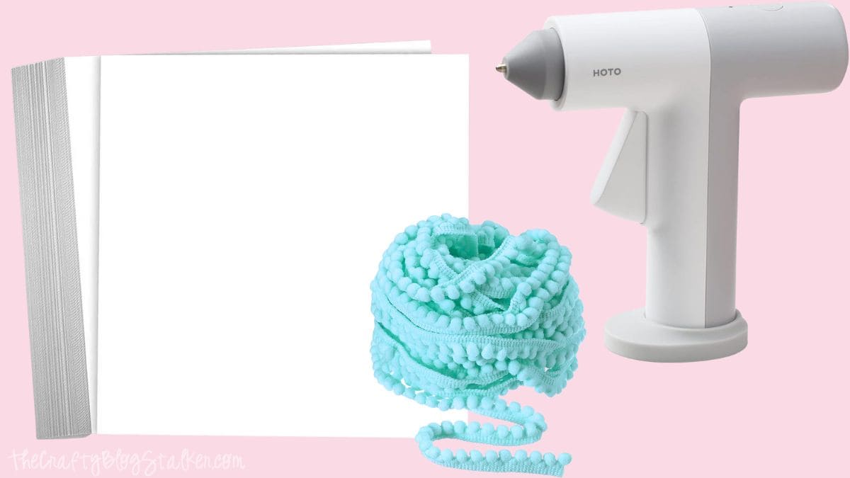 Craft supplies including white poster board, mint blue pom pom trim, and a white hot glue gun on a pink background.