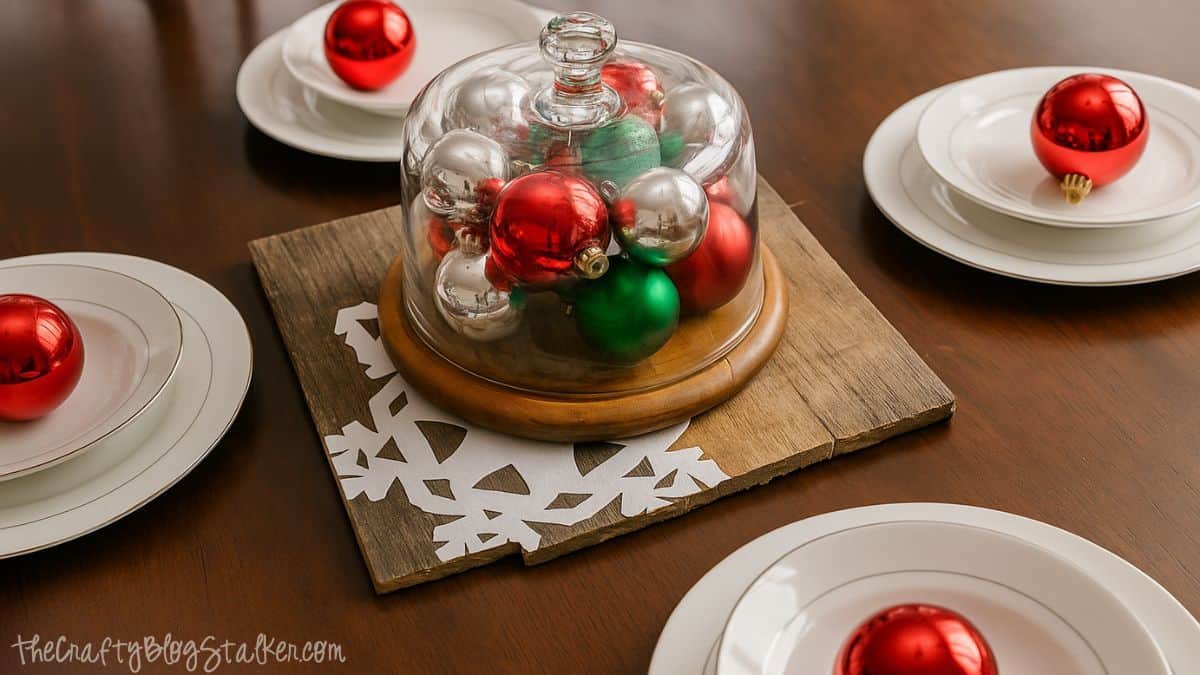Holiday table setting with a rustic wood snowflake charger holding a glass cloche filled with red, green, and silver ornaments surrounded by white plates.