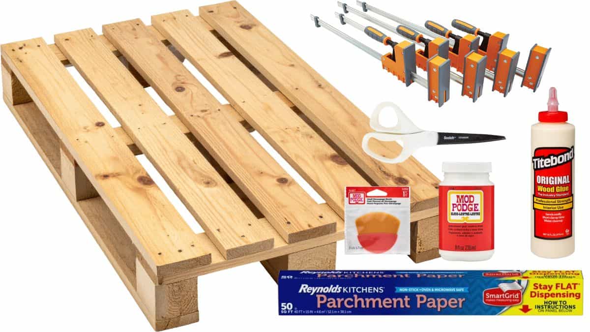 DIY pallet wood charger supplies including a wooden pallet, wood glue, clamps, scissors, Mod Podge, parchment paper, and a Mod Podge brush.