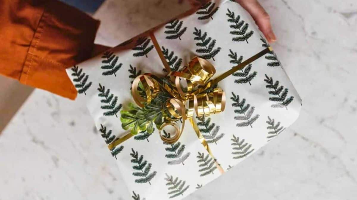 Gift wrapped in white evergreen-patterned paper with gold ribbon, curled bow, and greenery held over a marble surface.