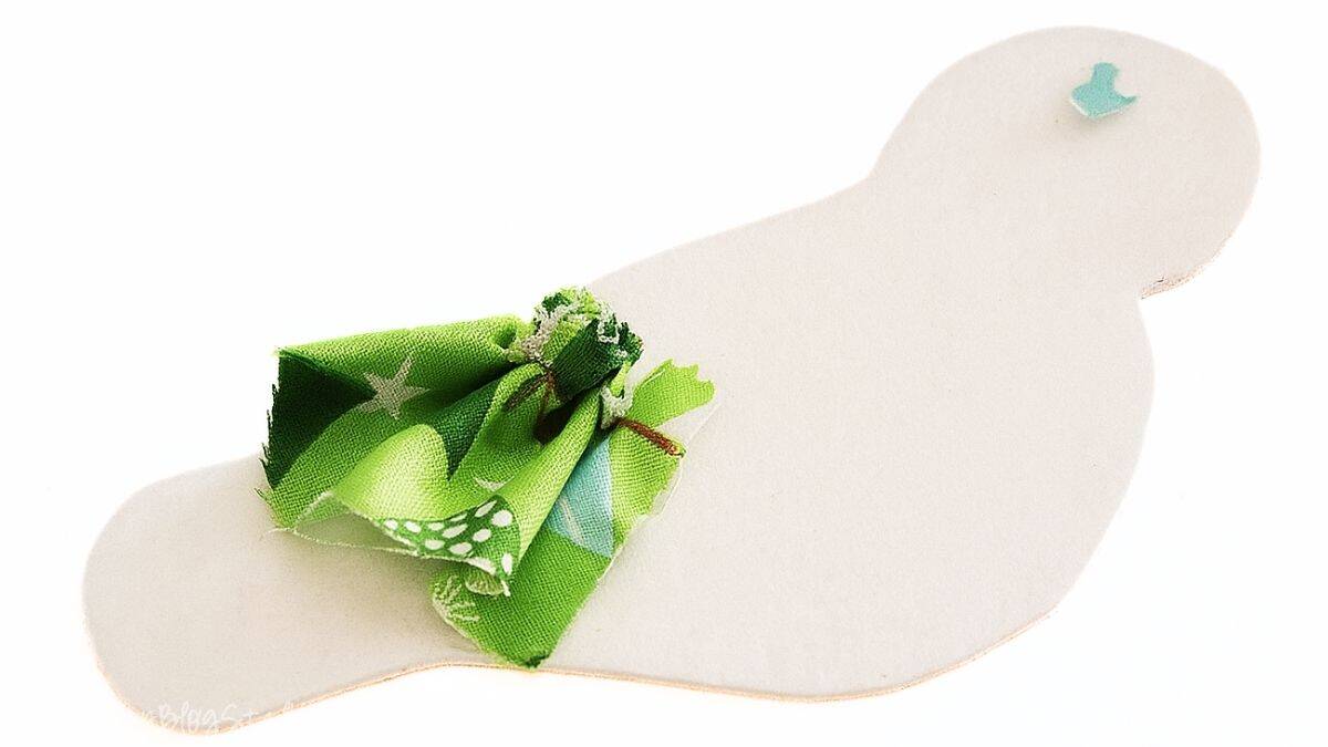 A white felt bird cutout with a small green fabric ruffle attached to its back, shown on a white background for a Christmas craft project.