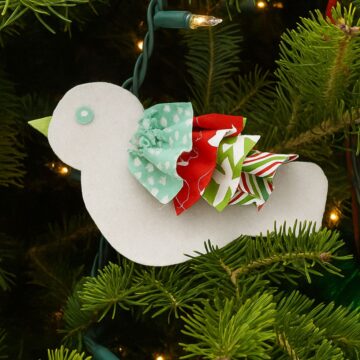 A white felt bird ornament with a green beak and layered red, green, and aqua ruffled fabric tail hanging on a Christmas tree with lights.