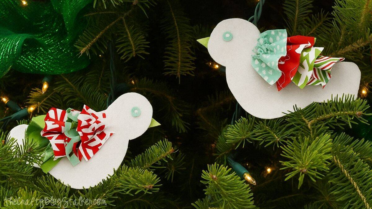 Two handmade bird ornaments with colorful fabric tails and button eyes hanging on a Christmas tree with lights.