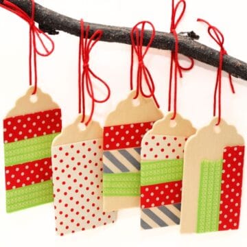 Five wooden tags decorated with patterned washi tape and hung from a branch using red string, creating simple handmade ornaments.