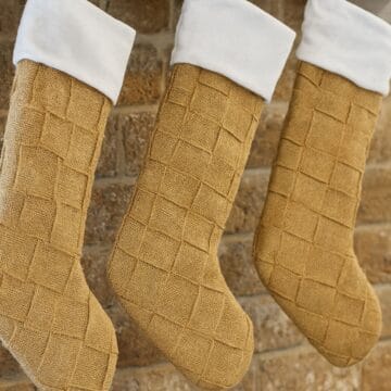 Three woven burlap Christmas stockings with fabric cuffs hanging side by side on a brick wall, showing a textured, handwoven pattern.