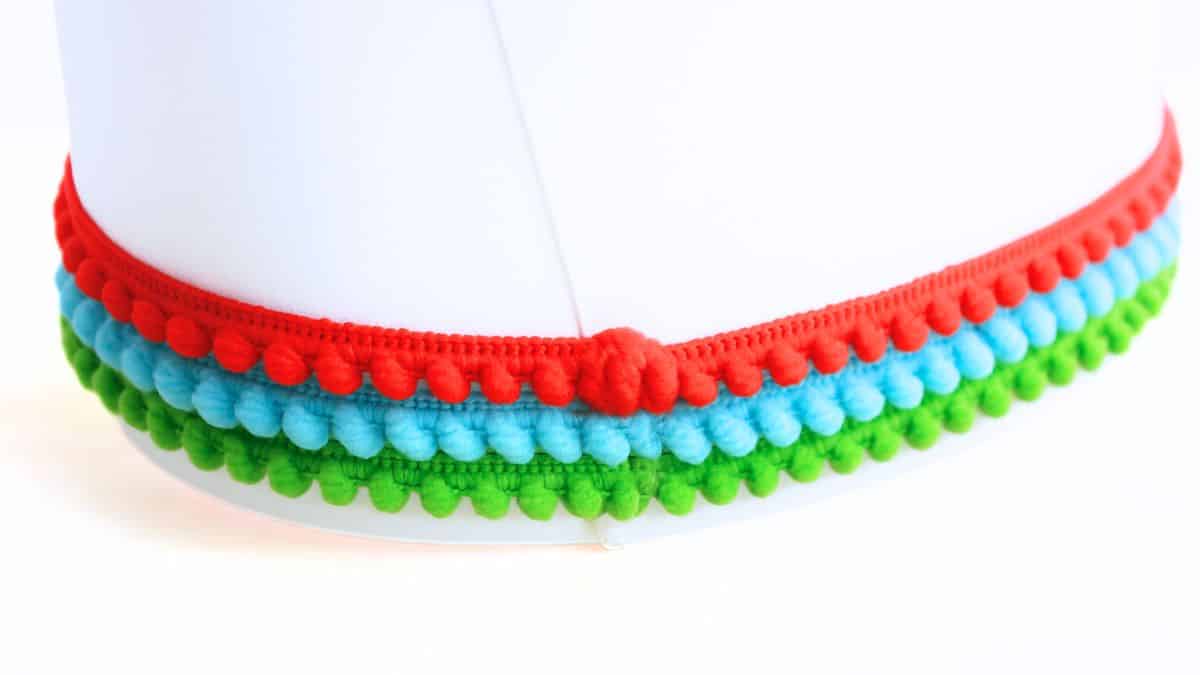 Close-up of red, blue, and green pom pom trim wrapped around the bottom edge of a white cone for a colorful Christmas craft.