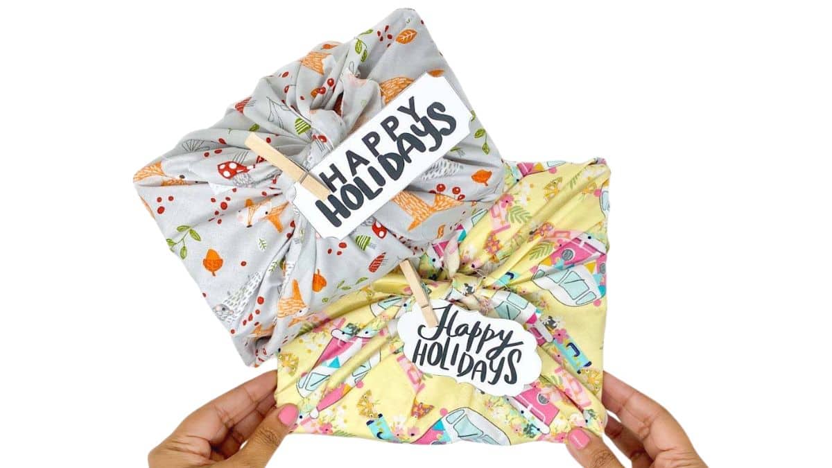 Two holiday gifts wrapped in colorful patterned fabric and finished with Happy Holidays gift tags clipped with wooden clothespins.
