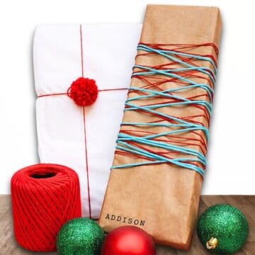 Two wrapped gifts on a wooden surface, one in kraft paper wrapped with yarn and stamped with a name, styled with yarn and decorative ornaments.