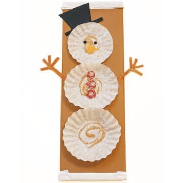 Coffee filter snowman craft with three stacked coffee filters, paper hat and carrot nose, pipe cleaner arms, and stamped details on a brown cardboard fabric bolt.