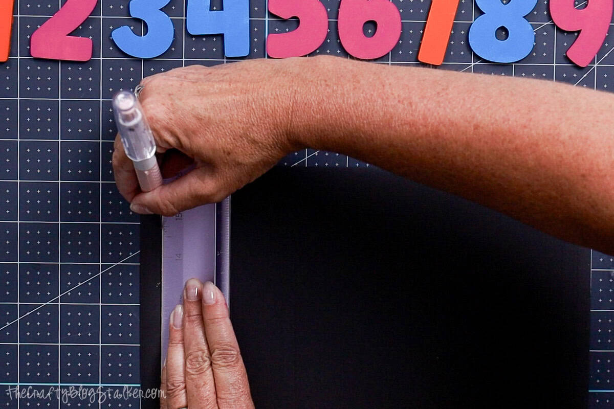 Person using a craft knife and ruler to cut black magnet sheets on a gridded cutting mat, with colorful painted wooden numbers arranged above.
