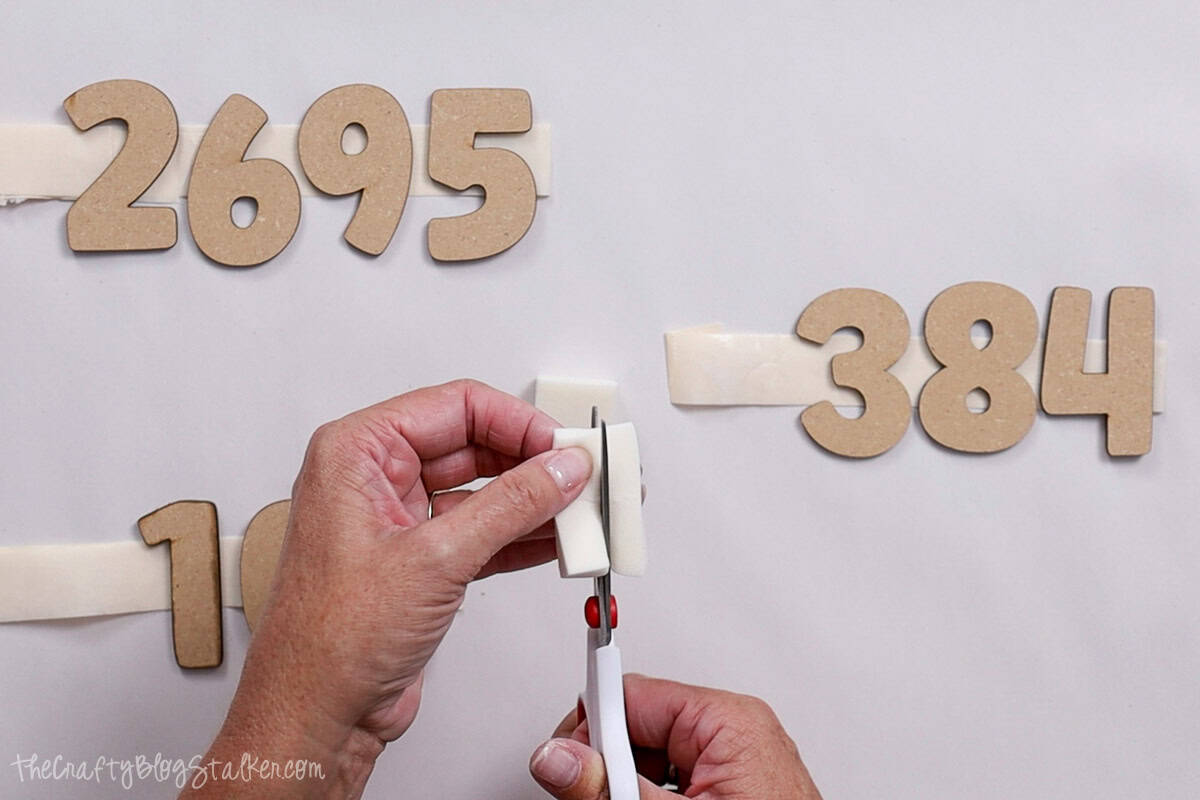Person using scissors to cut a makeup sponge while wooden numbers are taped to a surface in preparation for a DIY painting or stamping project.