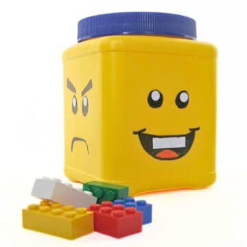 Repurposed plastic container decorated like a LEGO minifigure head used for LEGO storage, shown with loose LEGO bricks in front.