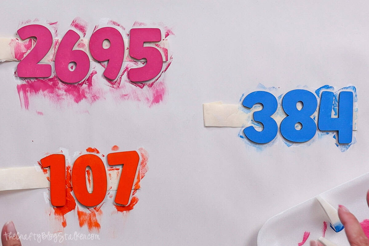 Painted wooden numbers in pink, orange, and blue drying on a taped surface, with paint smudges and sponge applicators nearby from a DIY craft project.