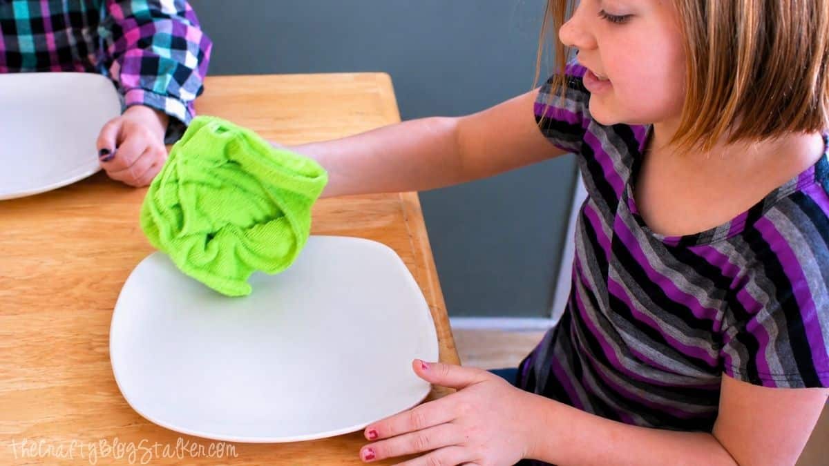 Girl wiping a white ceramic plate with a green cloth at a table, prepping the surface before decorating with Sharpie markers.