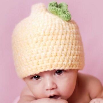 Baby wearing a yellow crochet hat with a small green leaf on top, lying on a pink background.