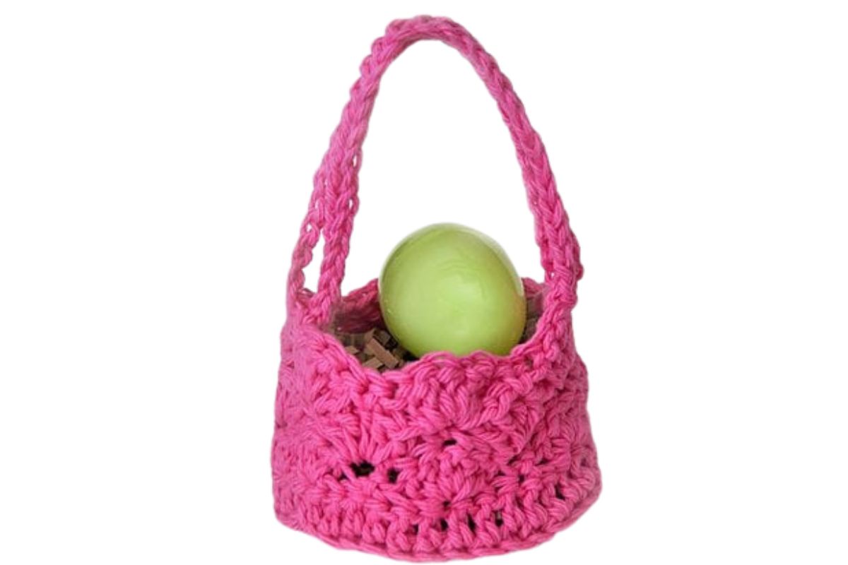 Pink crochet Easter basket with a scalloped edge and tall handle, holding a green plastic egg nestled in shredded paper.
