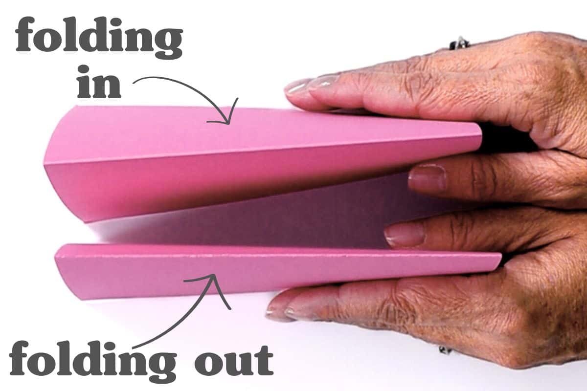 Hands holding folded pink paper with labels showing one fold going inward ("folding in") and another fold going outward ("folding out")