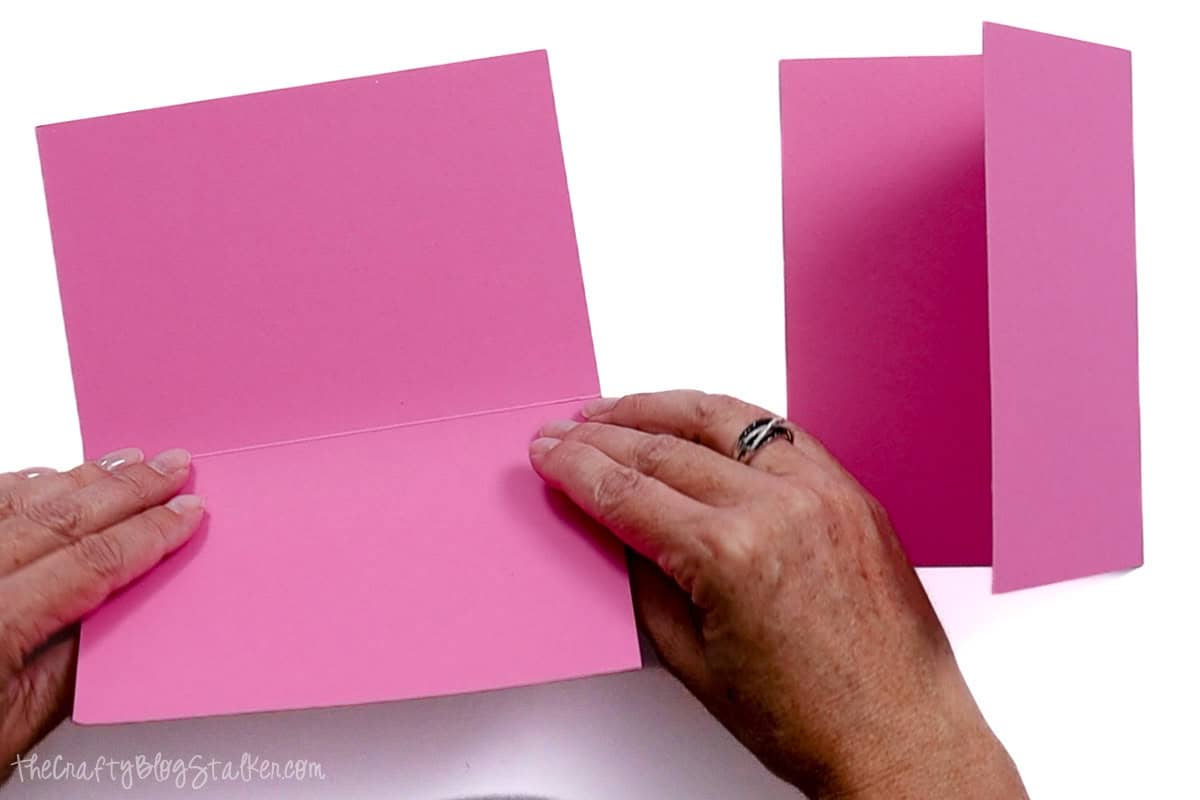 Hands folding pink cardstock outward along a scored line, with a second folded card standing upright in the background for a card-making project.
