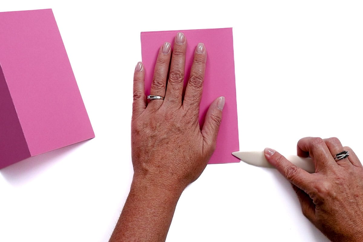 Hands folding pink cardstock using a bone folder on a white surface, with one card already folded and set aside.