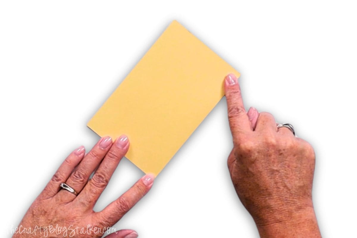 Hands folding a piece of yellow cardstock along a scored line, demonstrating a clean and precise paper fold for a crafting or cardmaking project.