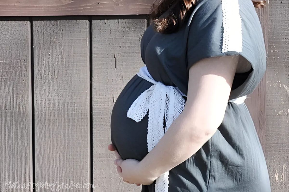 Side view of a pregnant woman wearing a dark gray maternity dress with lace accents and a white lace ribbon tied around her belly, standing in front of a wooden fence.