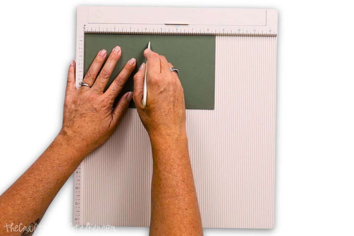 Hands scoring green cardstock on a scoring board using a bone folder, preparing the paper for a precise and clean fold in a crafting project.
