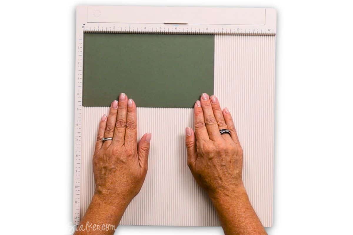 Hands positioning green cardstock on a scoring board, aligning it for accurate measurement and scoring in preparation for folding a paper craft project.