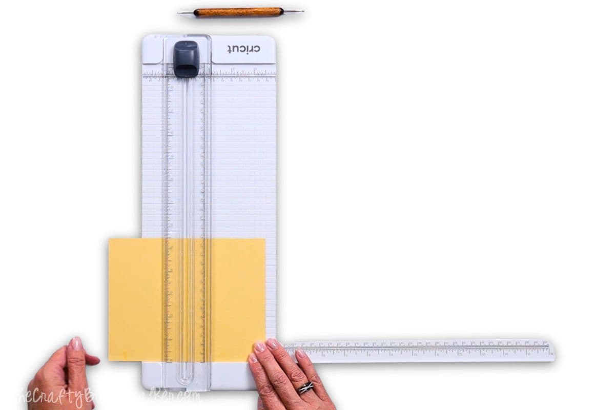 Hands positioning yellow cardstock on a Cricut paper trimmer, preparing to score using the stylus tool shown above on a clean white background.