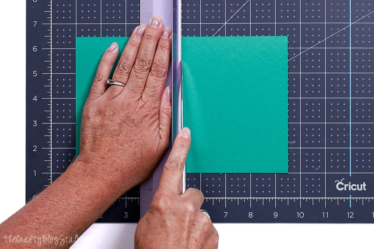 A hand using a butter knife along a Cricut paper trimmer edge to score a piece of teal cardstock, positioned on a Cricut cutting mat with grid lines.