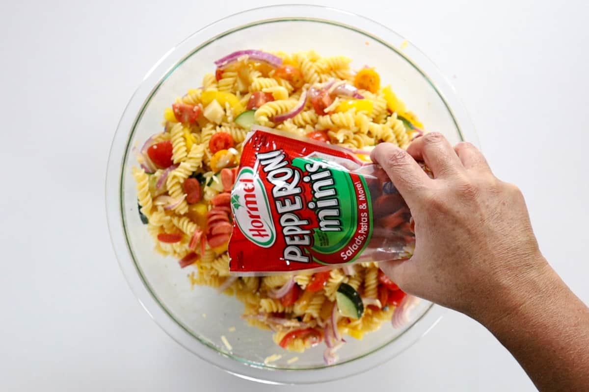 Pouring pepperoni minis into a glass bowl filled with gluten-free rotini pasta, tomatoes, cucumber, bell pepper, red onion, mozzarella cheese, and parmesan cheese.